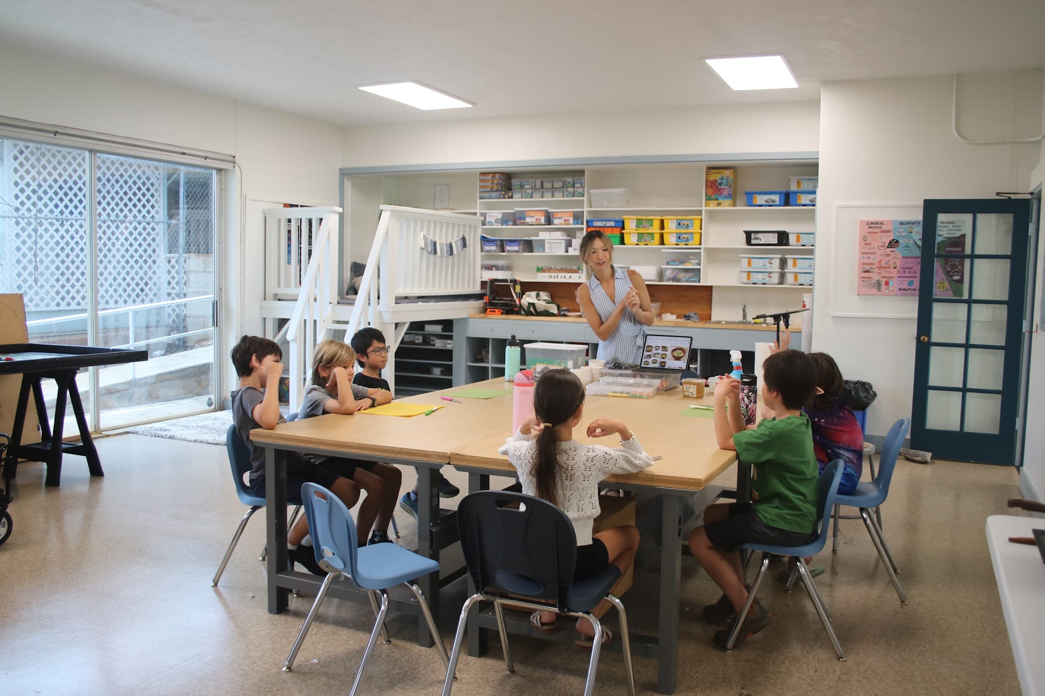 A wide art room: seven scholars around a large wooden table with coloured paper and a pencil case on it, a teacher standing at the back demonstrating with both hands, storage shelves of bins and paper stock along the back wall.