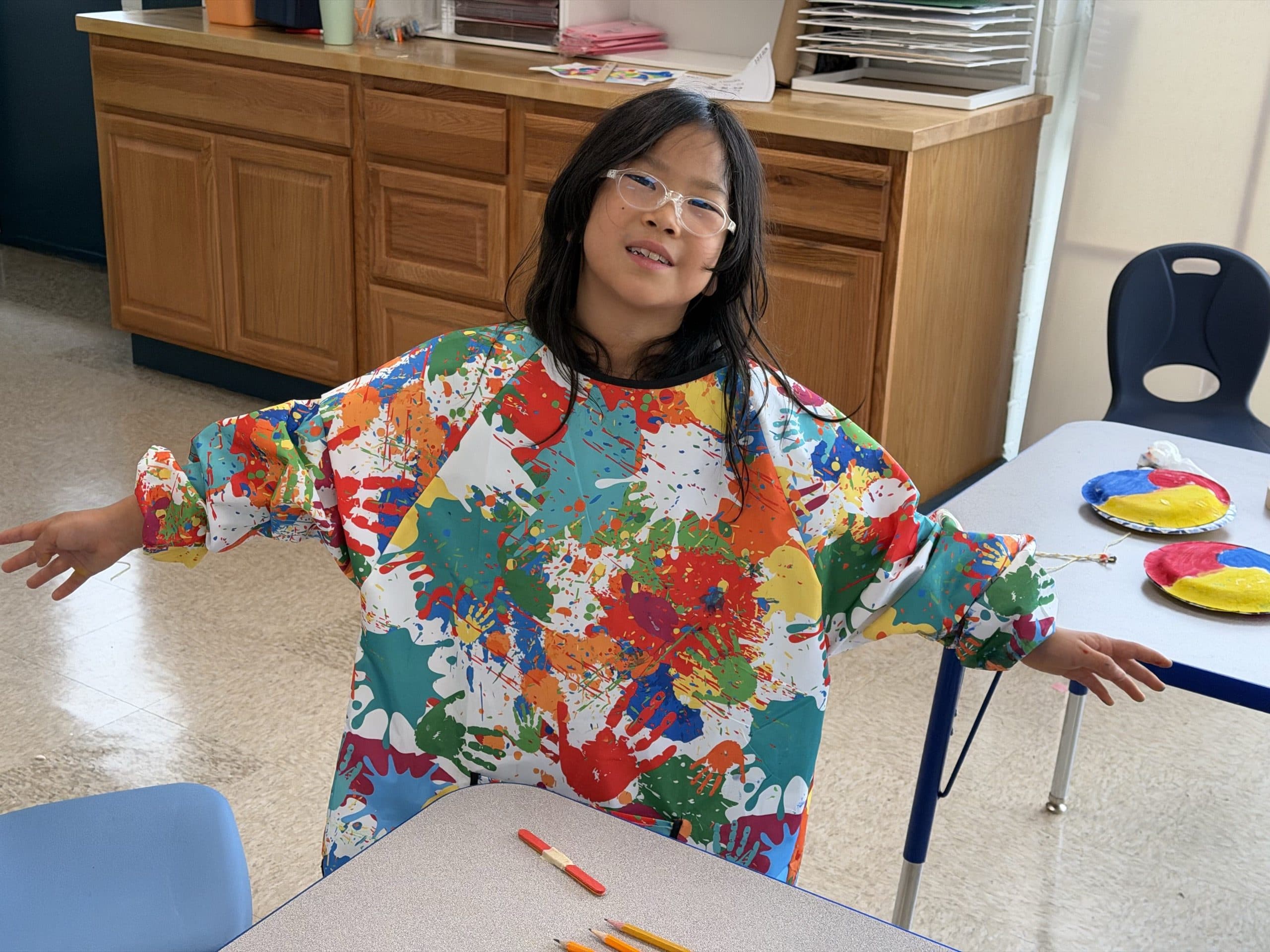 A scholar in the art room showing off a paint-splattered smock with her arms held out wide, grinning, pencils on the table in front of her and two painted paper plates in red, yellow and blue on the table behind.
