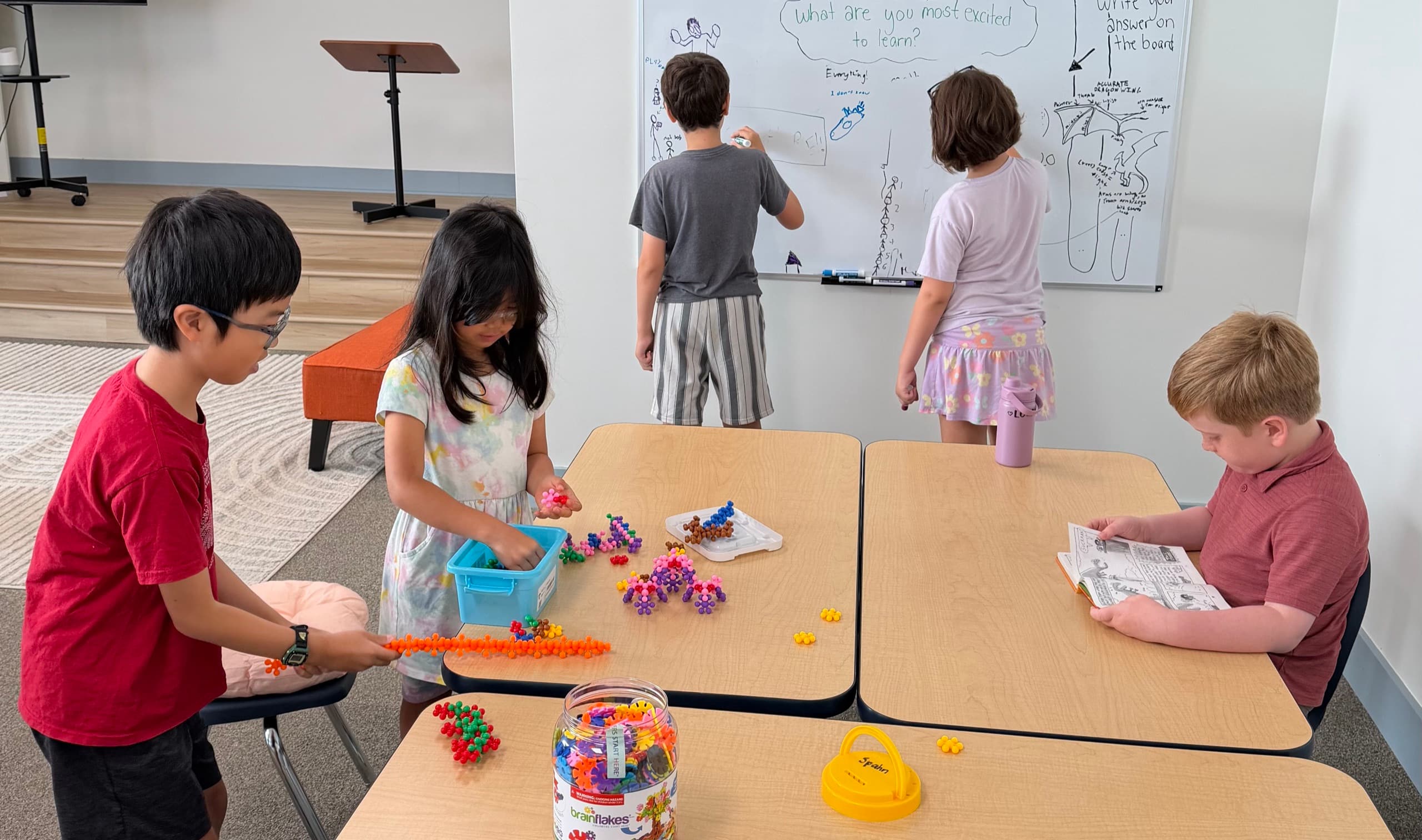 A classroom on the first morning: two scholars writing answers on a whiteboard headed “Happy 1st Day of School”, three more at tables behind them building with plastic construction pieces and reading.