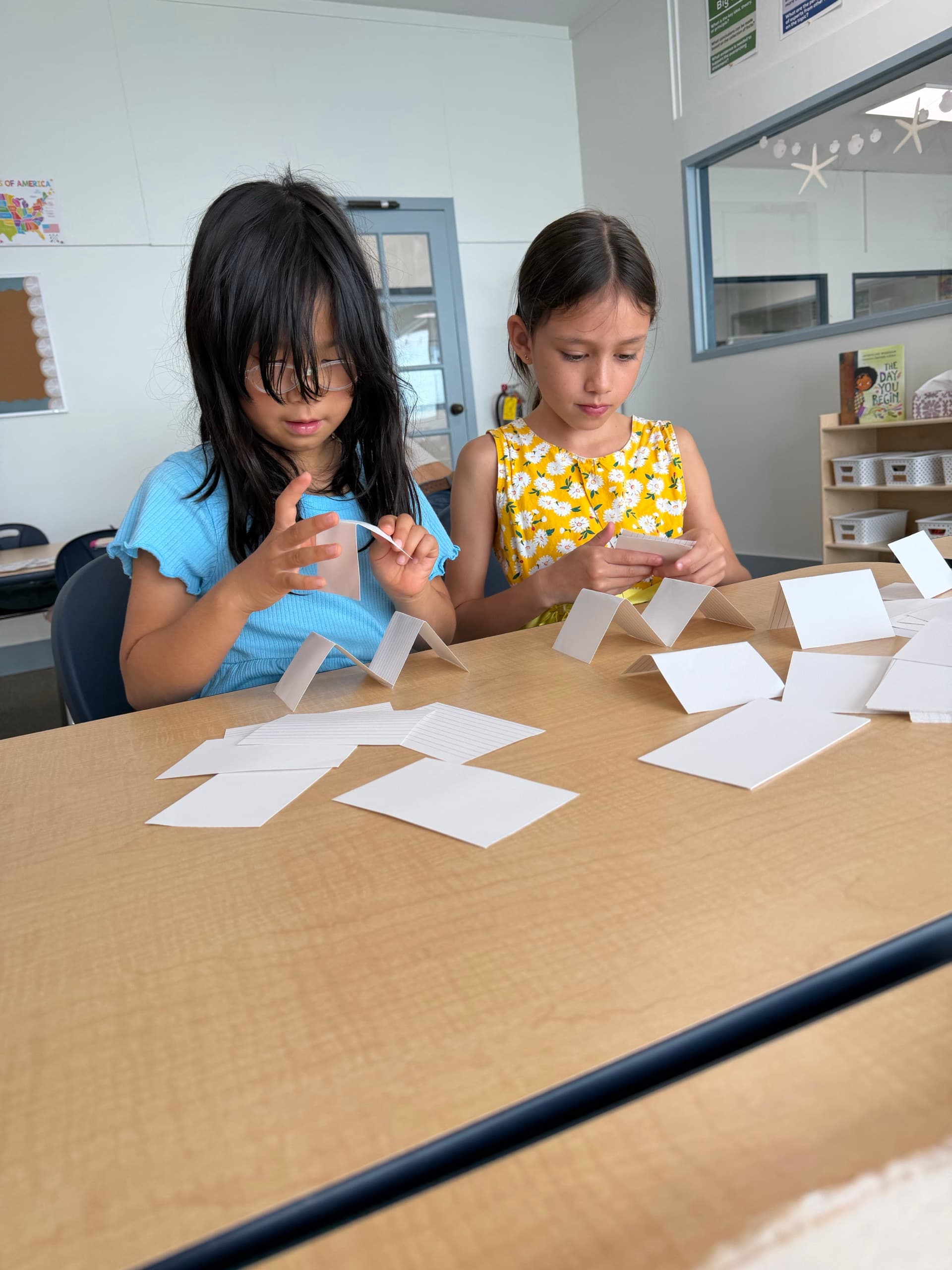 Two scholars at a wooden table sorting blank index cards into standing rows, each holding a card and reading it, a classroom map and a shelf of labelled bins behind them.