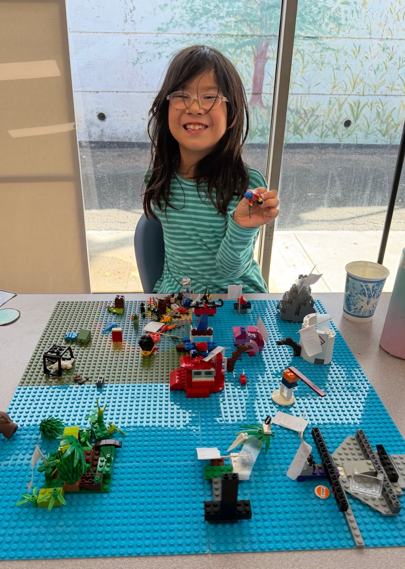 A scholar at a table of blue Lego baseplates covered in a built-out scene, holding one small figure up to the camera and grinning.