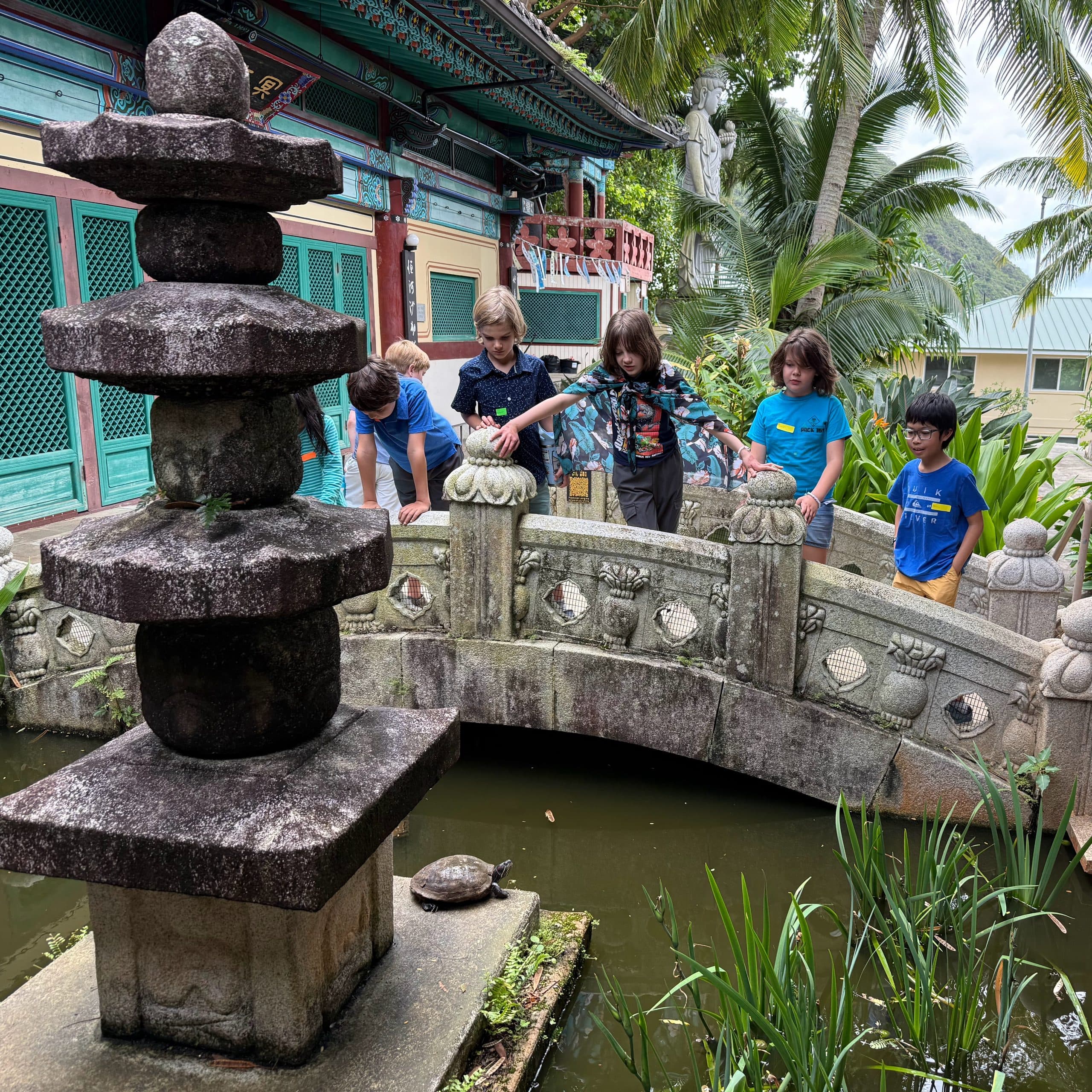A field trip to Mu-Ryang-Sa, the Korean Buddhist temple in Pālolo: scholars leaning over the carved stone rail of a small arched bridge above a pond, a weathered stone pagoda in the foreground, the temple's painted eaves and a standing statue among palms behind them.
