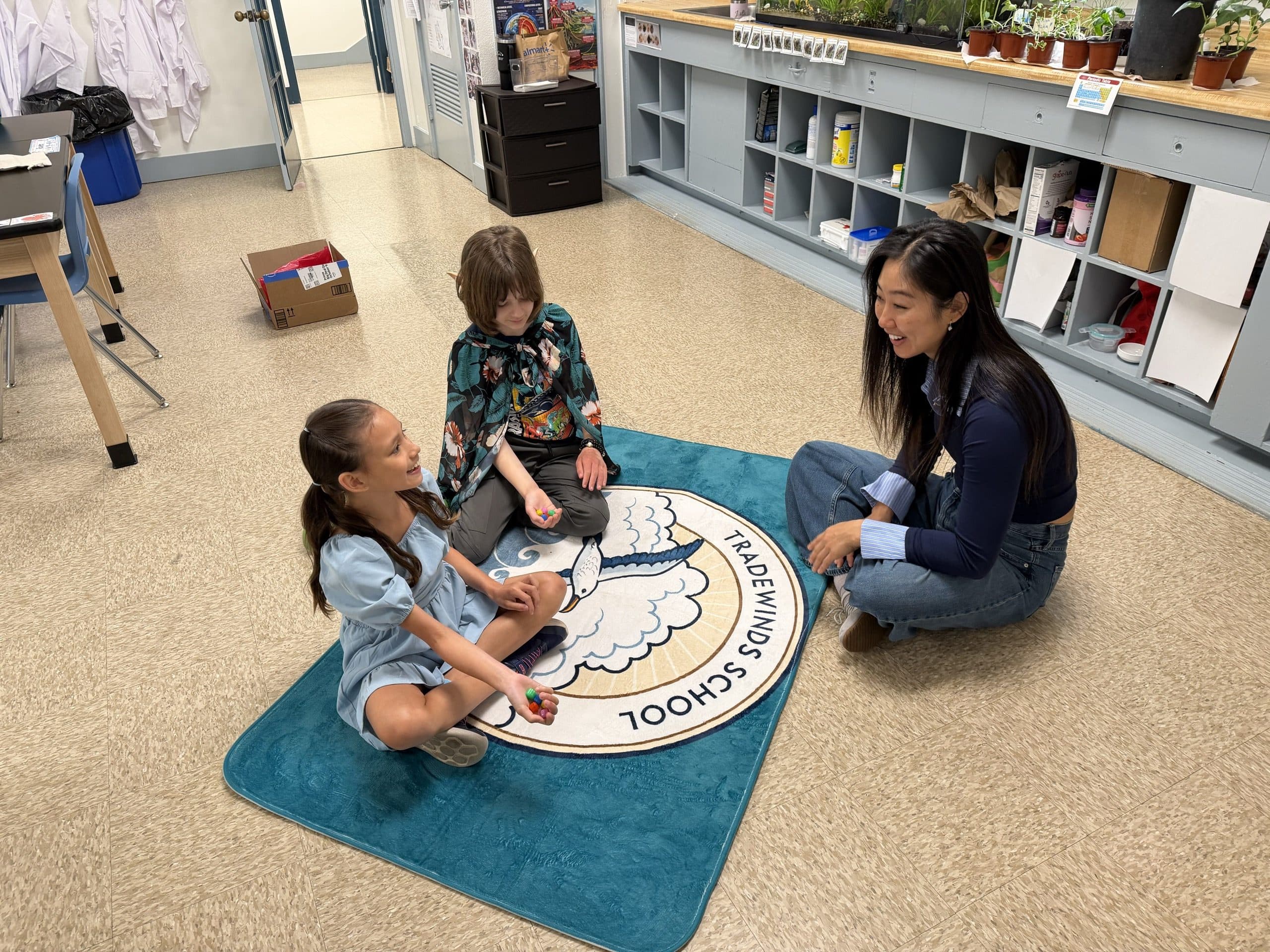 Two scholars sitting cross-legged on the albatross rug playing a game with small coloured pieces, a teacher sitting on the floor with them and laughing along, cubbies and potted plants behind.