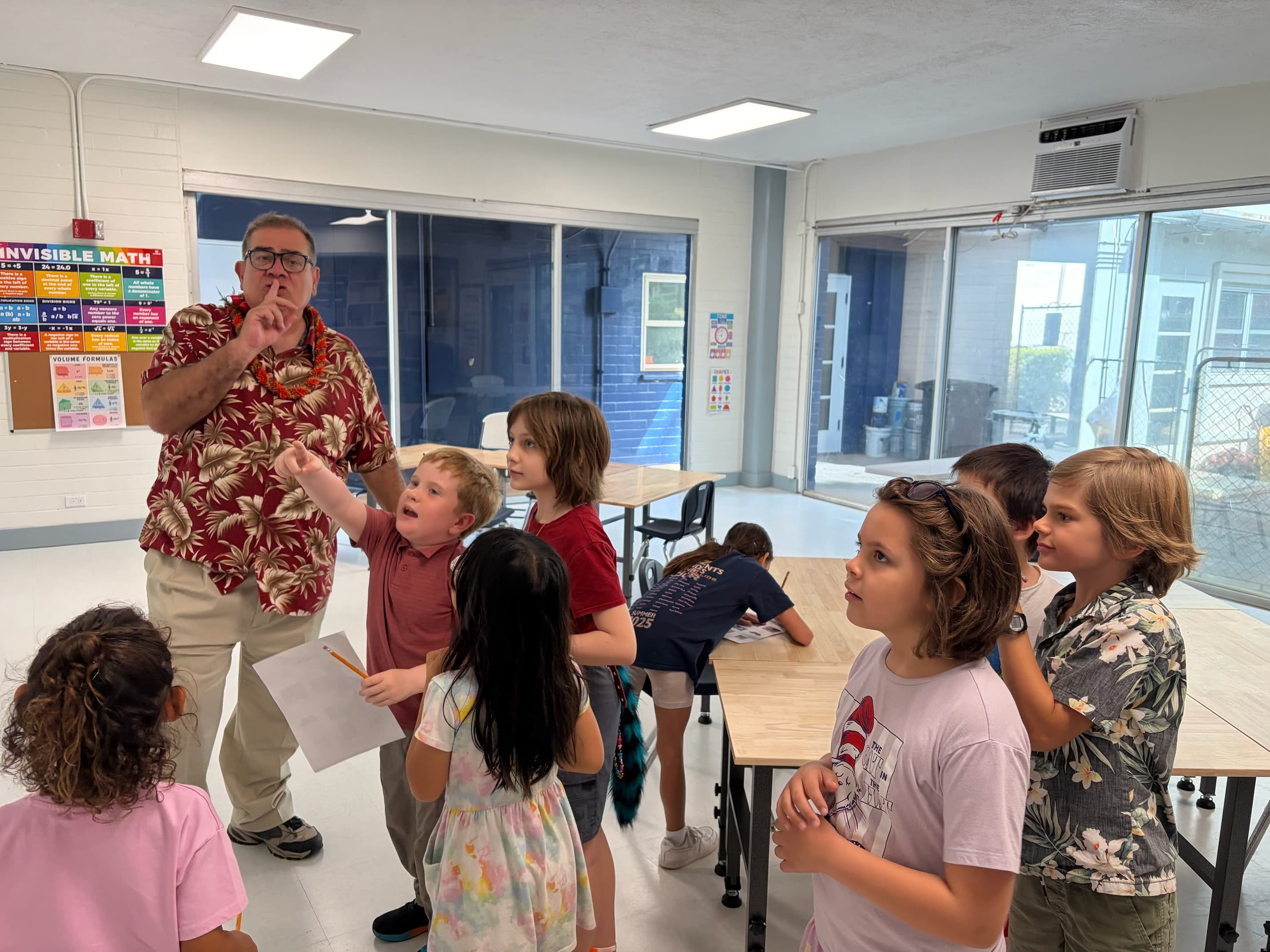 A scavenger-hunt riddle: an adult with a finger to his lips holding a clue sheet, eight scholars pressed in around him, one shouting an answer with an arm out, one holding the clue paper, one still writing at a table behind.