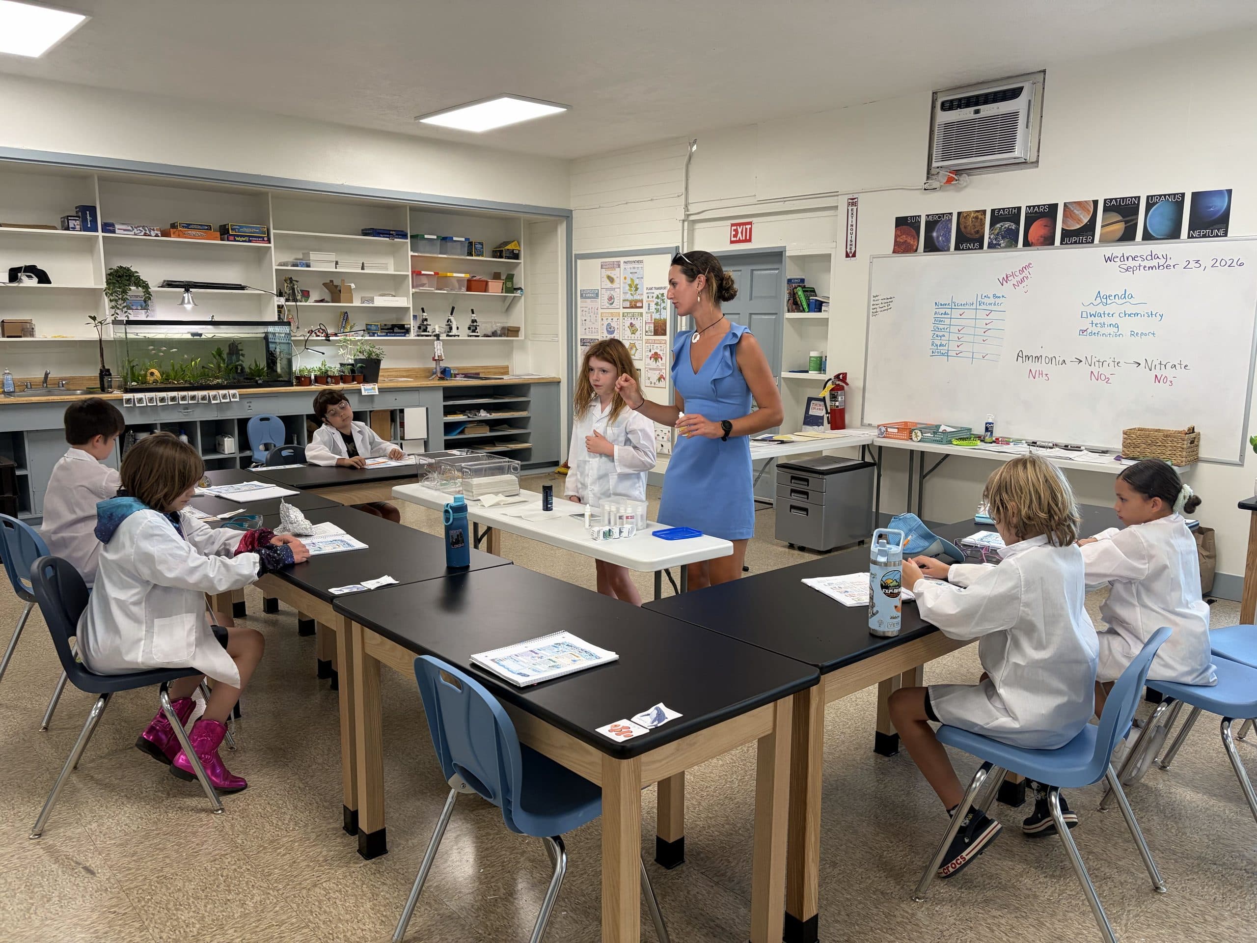 A science class at black lab benches: scholars in white lab coats working through printed sheets and small cards, a teacher standing at the front talking with one scholar beside a tray of beakers, and a whiteboard agenda on ammonia, nitrite and nitrate under a row of planet posters.