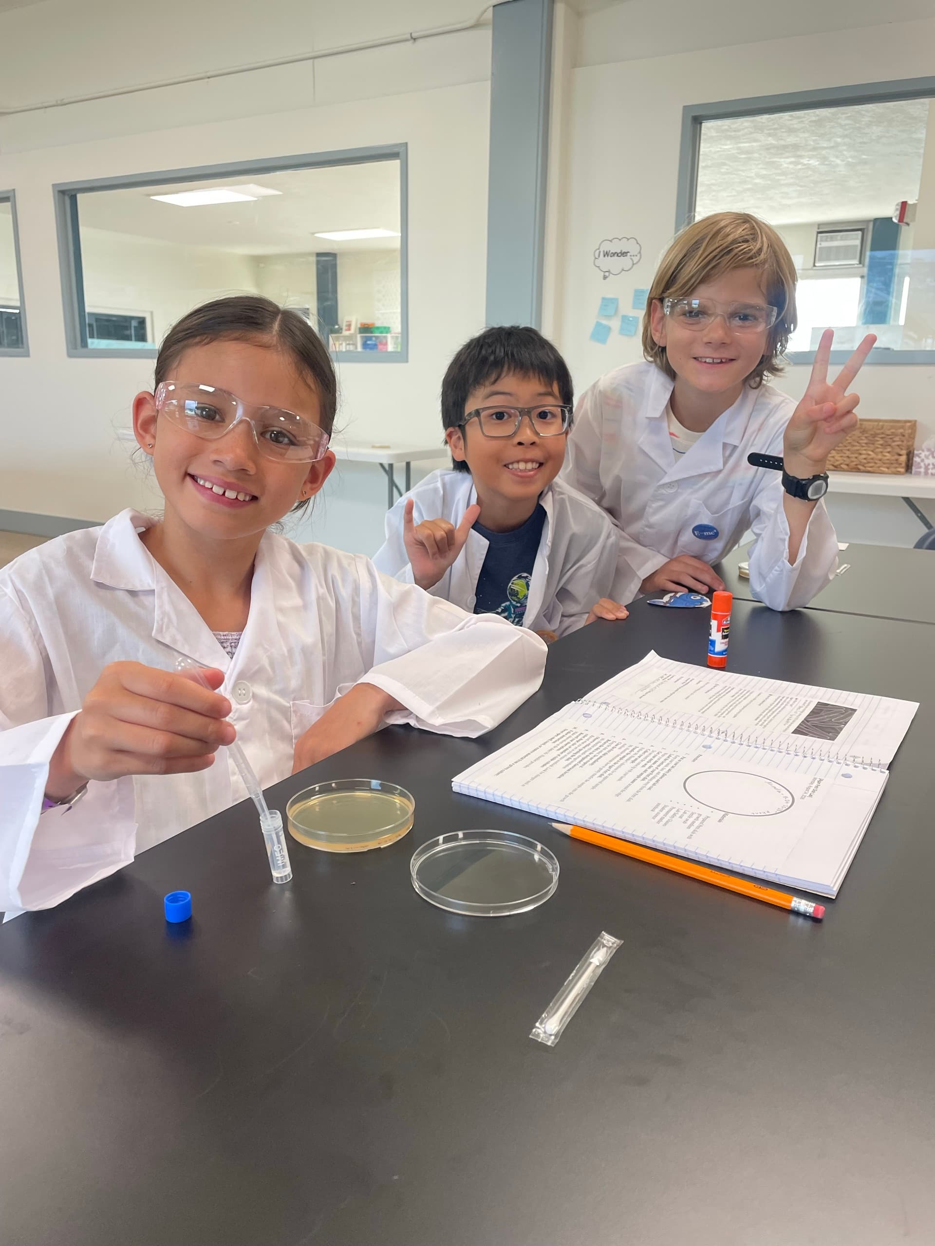 Three scholars in white lab coats and safety glasses at a black science bench, one holding a pipette over an agar plate, a second plate and a lab notebook open beside them.