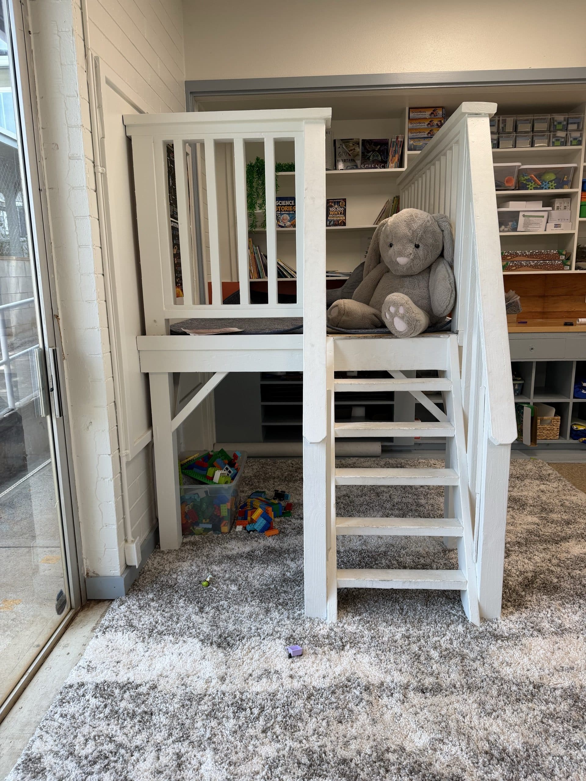 A soft space in a classroom: a white wooden loft with a short staircase, a large grey plush rabbit sitting on the cushioned deck, a bin of building bricks and a few loose pieces on the shaggy grey rug underneath, shelves of books and labelled bins behind.
