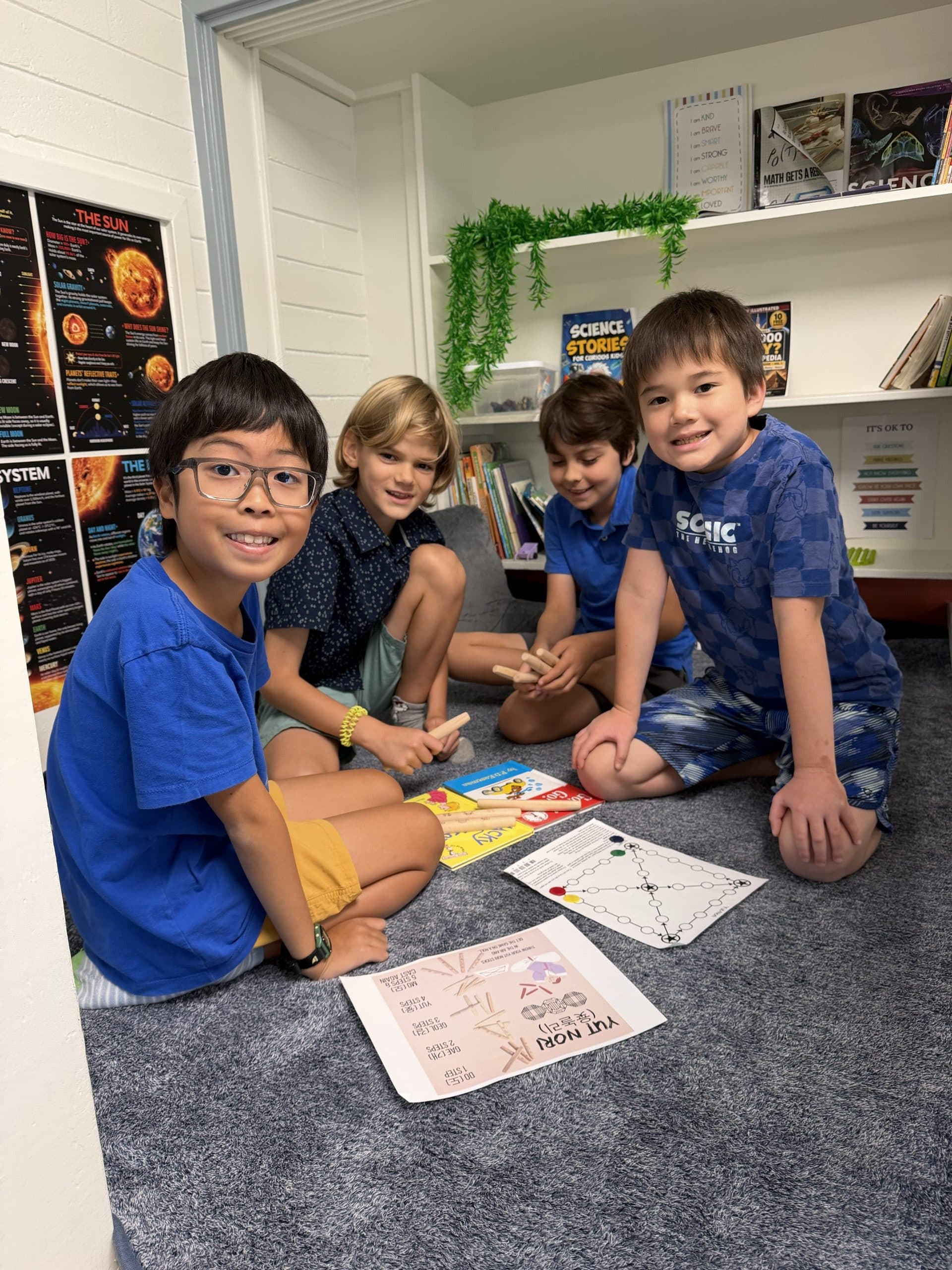 Four scholars kneeling on the carpet under the reading loft, grinning at the camera over a game of yut nori: the printed rules sheet, a board drawn on paper and the wooden sticks between them, shelves of science books behind.