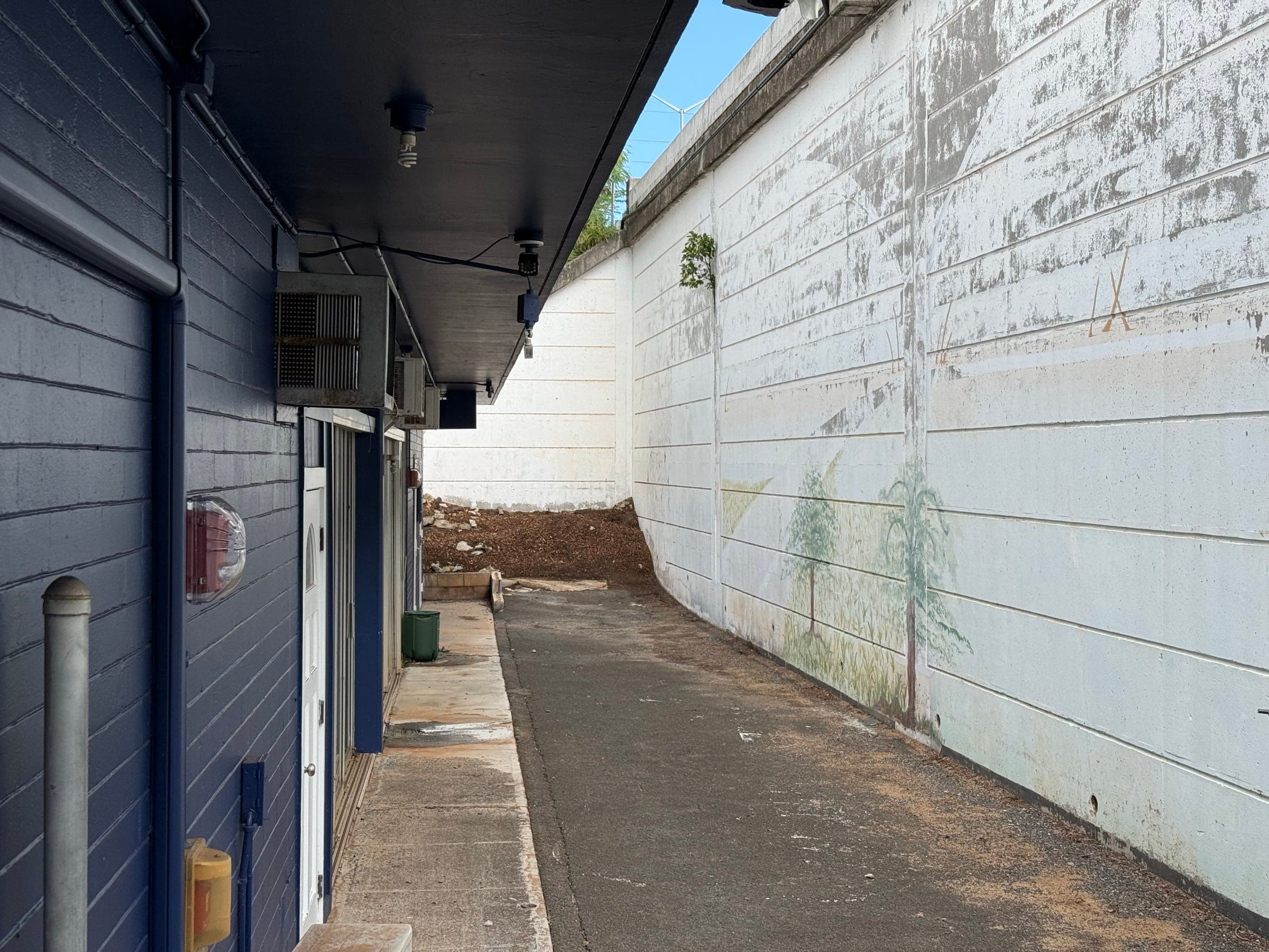 The alley behind the main building today: a strip of asphalt between the navy-painted classrooms and a tall white retaining wall, with a pile of dirt at the far end.