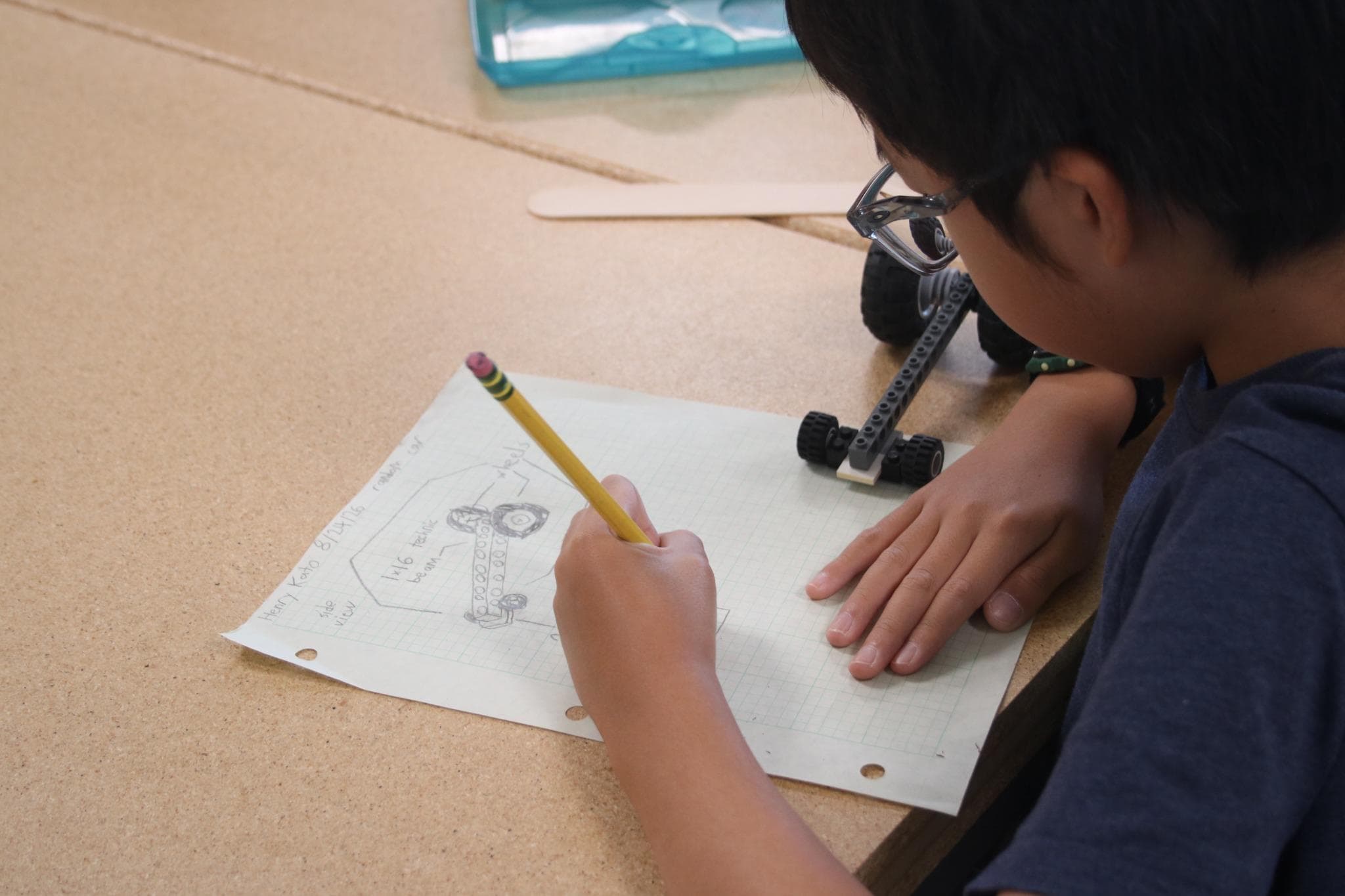 A scholar with the pencil down on his own labelled sketch of a wheeled build, the build itself sitting on the graph paper beside the drawing.