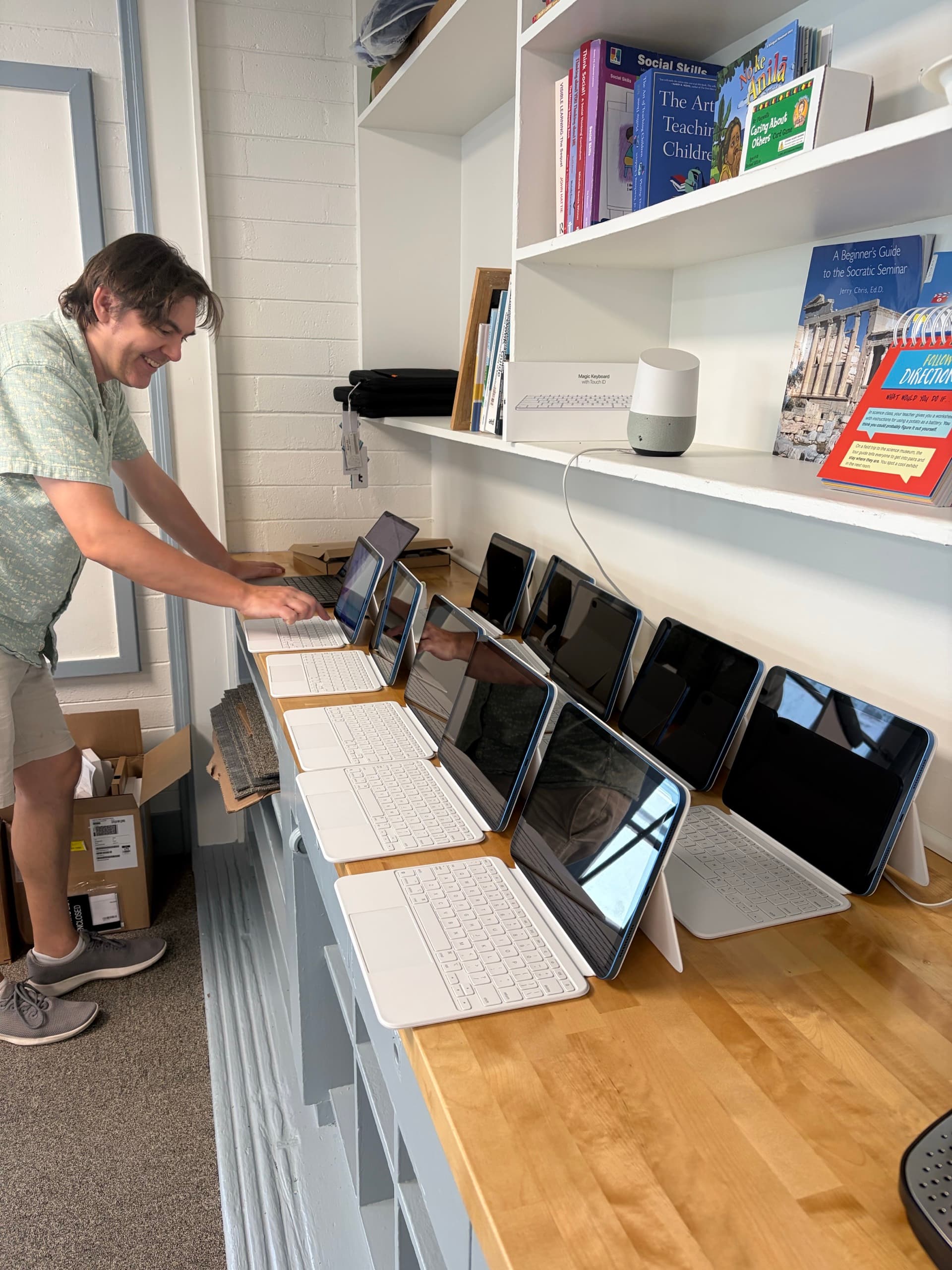 A row of BunnyPads stood up on a bench with their keyboard covers open, an adult leaning in to set up the one at the end, shelves of teaching books above them.