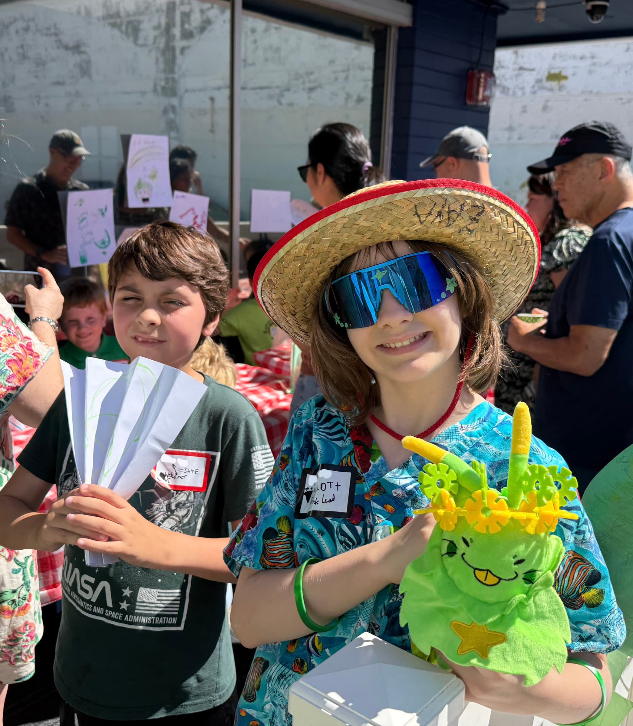 Two scholars at the limeade stand, close in and grinning, one in a straw hat and mirrored sunglasses holding a green plush mascot, the other holding a fan of hand-drawn flyers, hand-lettered signs and families behind them.