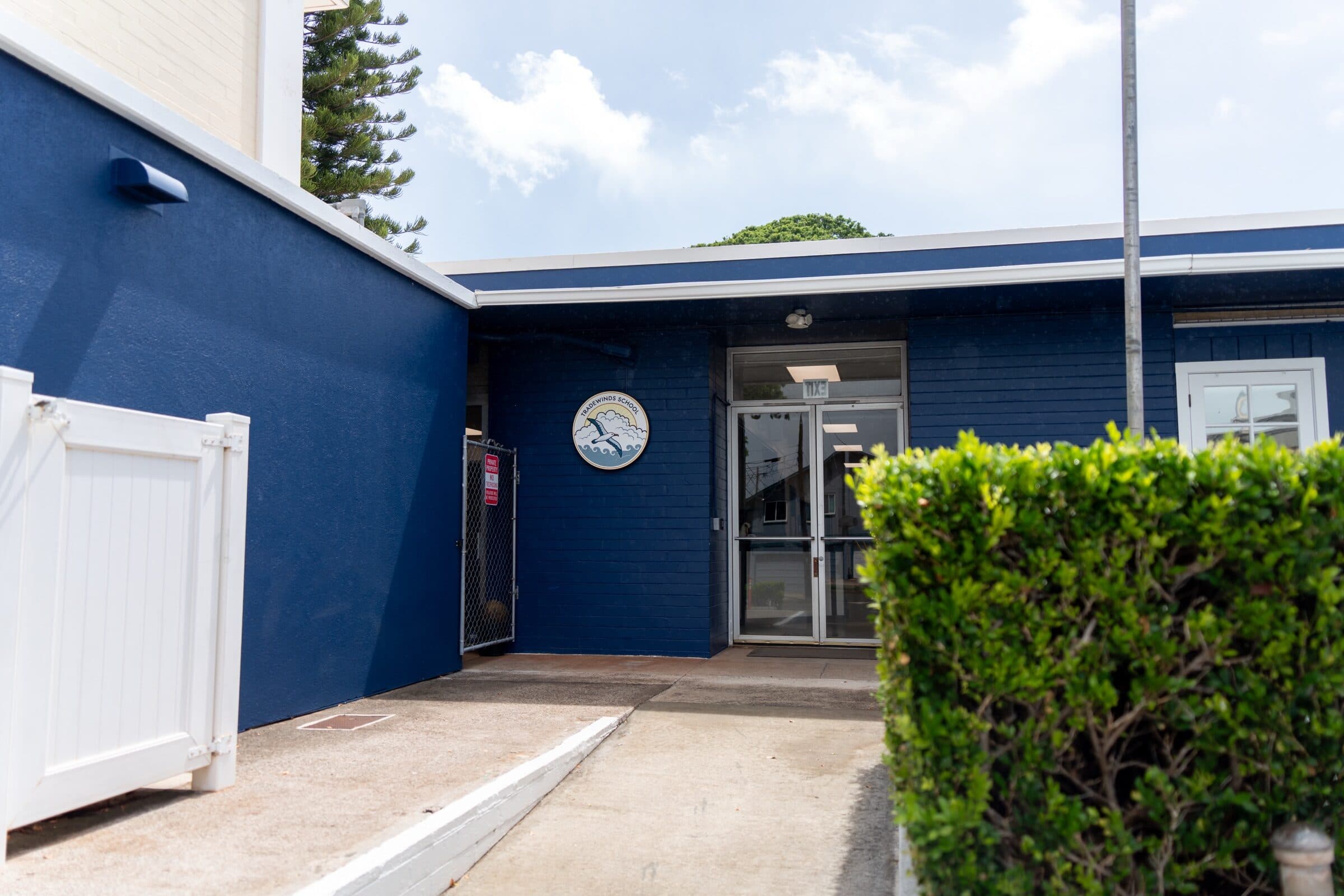 The entrance to the blue main building: glass double doors under a white fascia, the albatross seal on the navy wall beside them, a white gate on the left and a clipped hedge on the right.