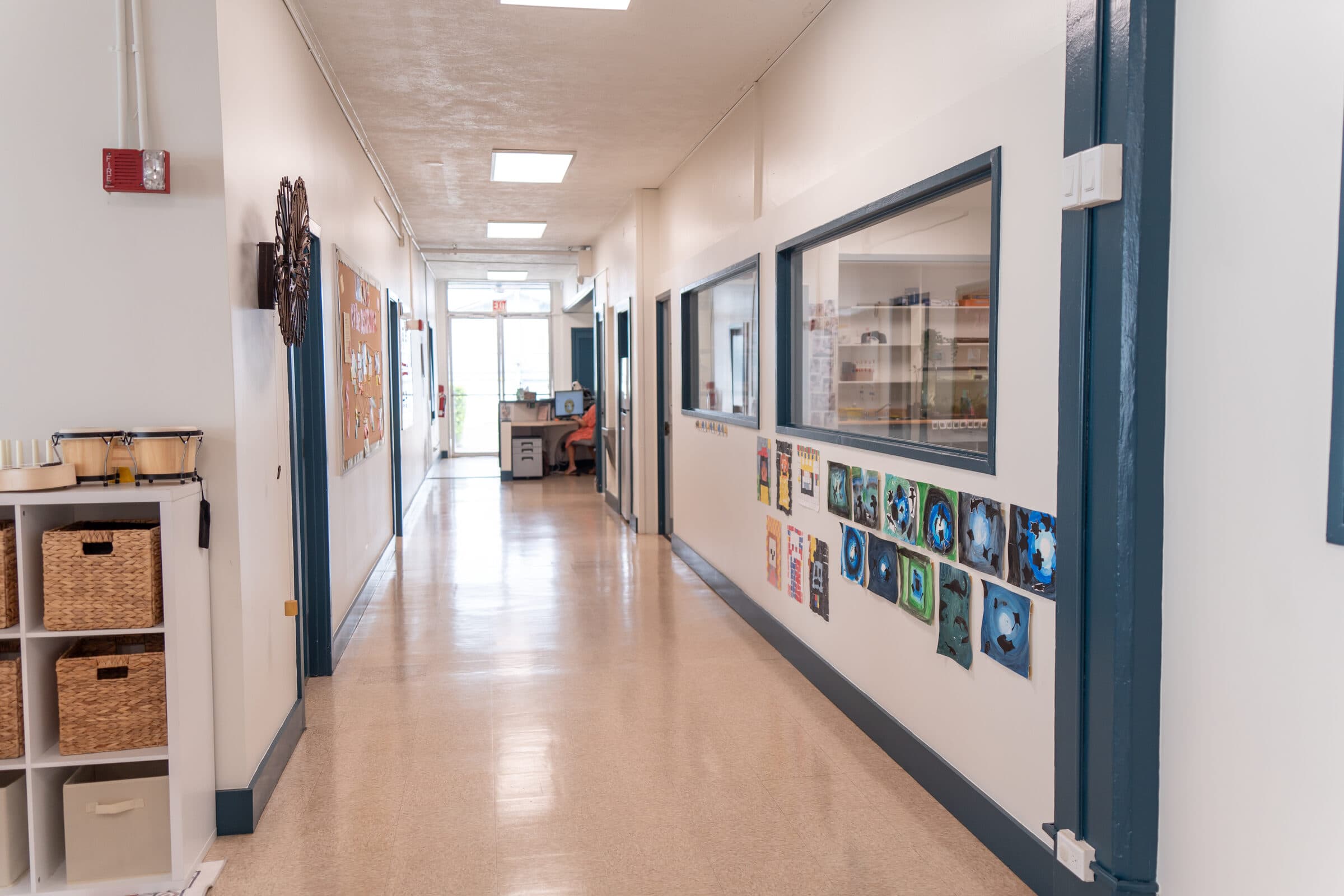 The long corridor in the main building: a polished floor running to glass doors at the far end, navy door frames, scholars' paintings taped below an interior window on the right, a cubby shelf with woven baskets and hand drums on the left.