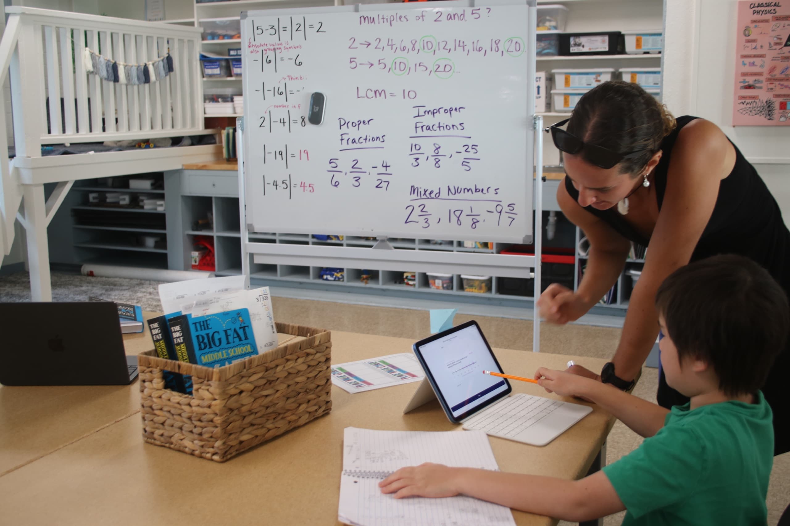 A scholar working at a table with a tablet open on a stand and a spiral notebook in front of him, pencil in hand, a teacher leaning in beside him to look at the screen, a whiteboard of fraction work behind them.