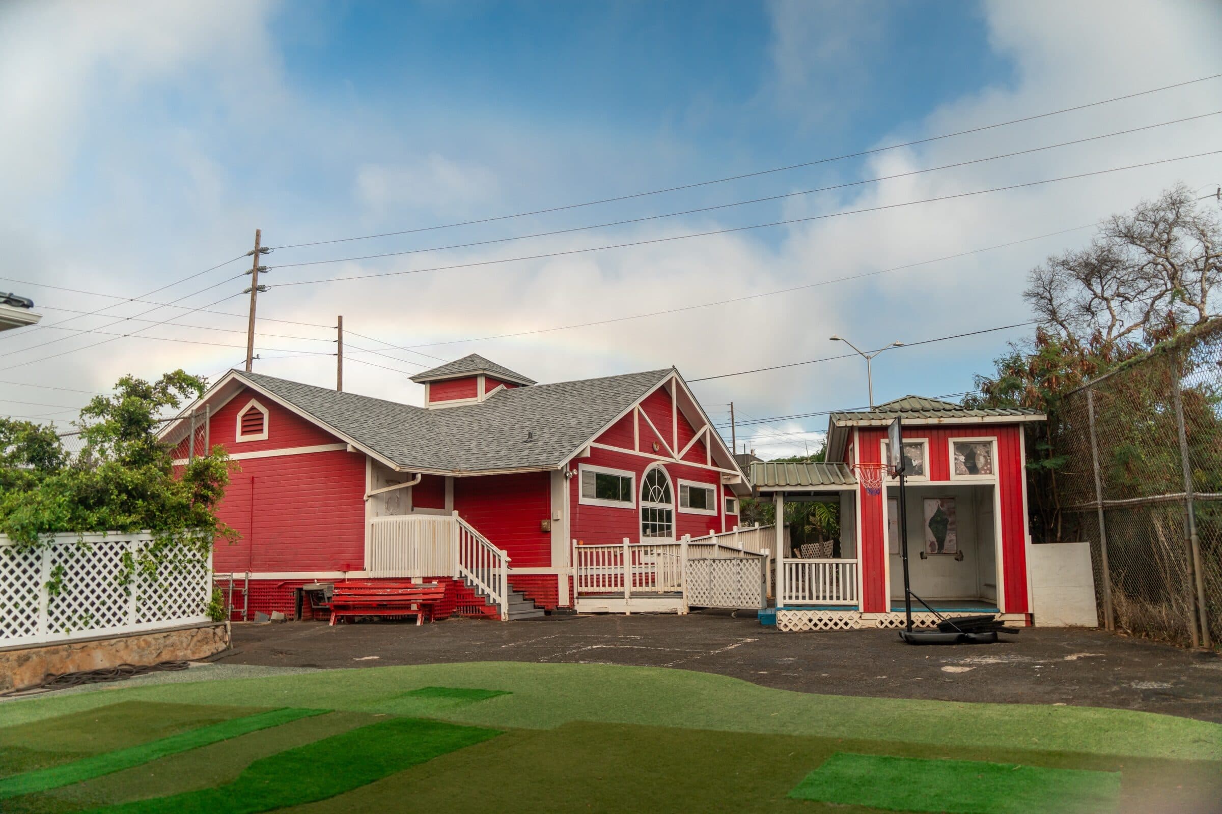 The red schoolhouse seen across the yard: white railings up the front steps, an arched window in the gable, the small red cottage with a basketball hoop beside it and turf in the foreground.