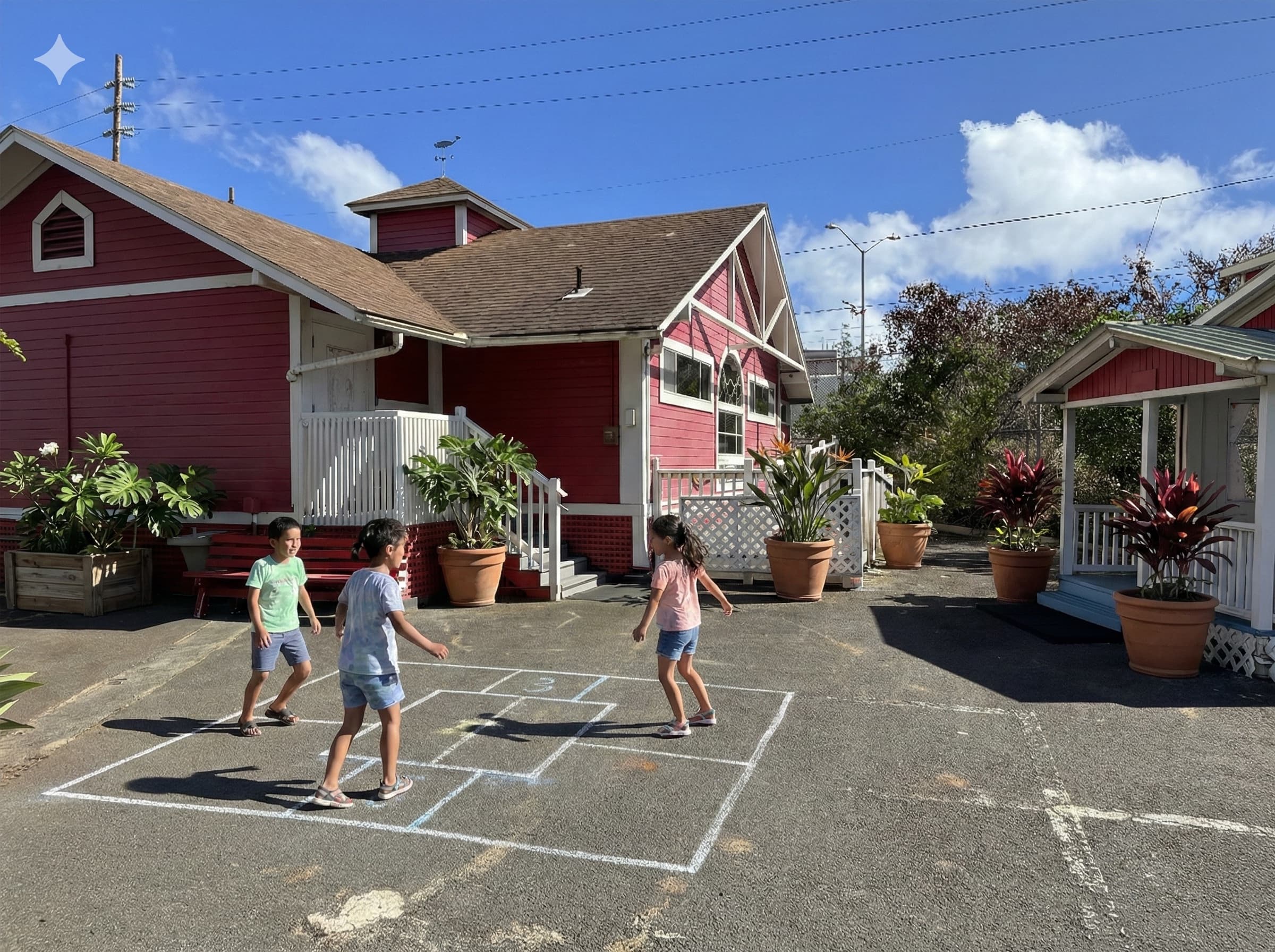The red schoolhouse and the smaller red cottage beside it, planted containers along both buildings and a hopscotch grid chalked on the yard.